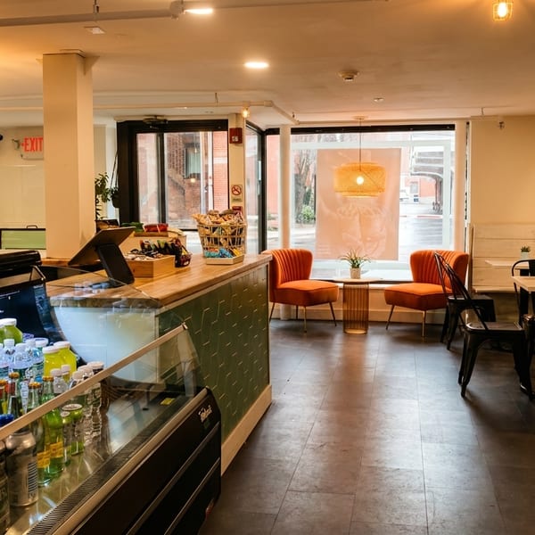 Modern cafe interior with a refrigerated display case, counter, and seating area with orange chairs.