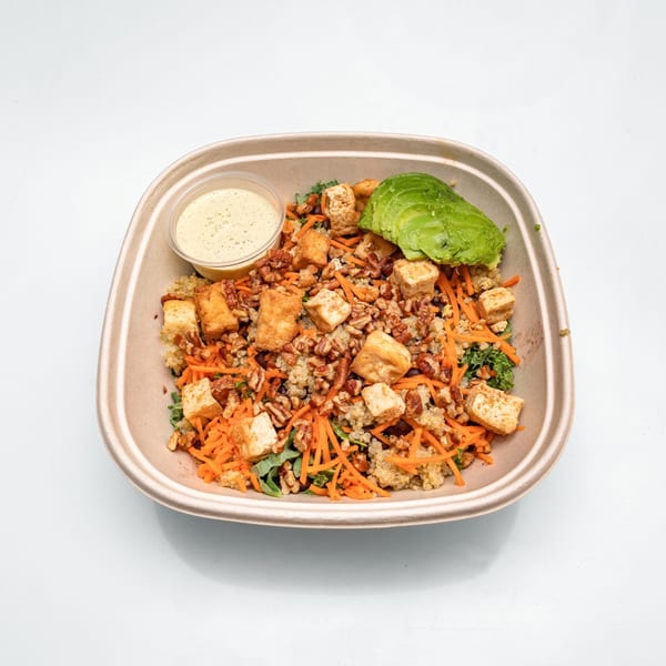 A healthy salad in a brown paper bowl with tofu, carrots, avocado, and dressing.