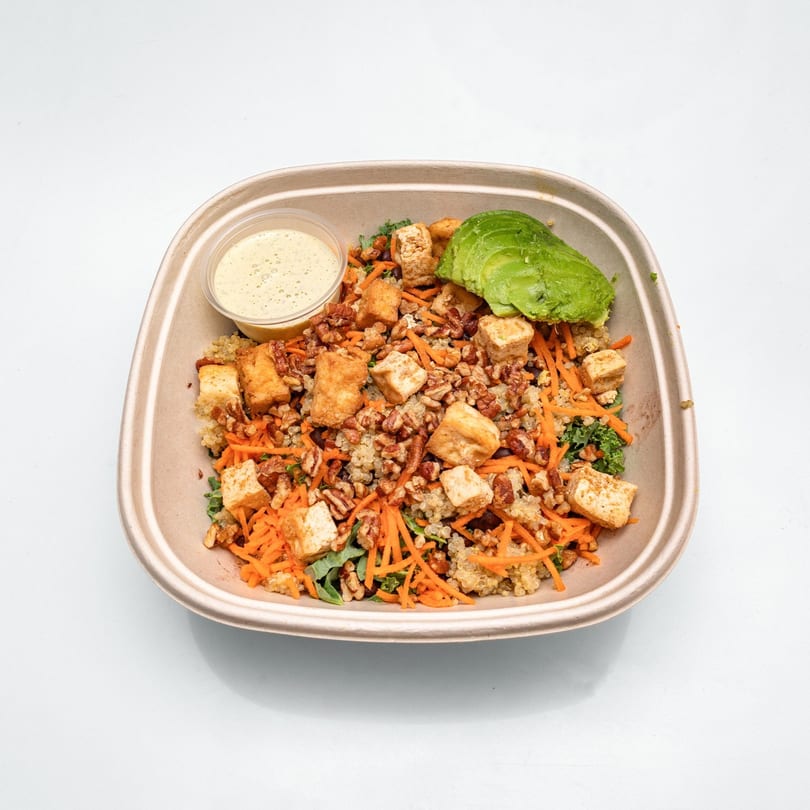 A healthy salad in a brown paper bowl with tofu, carrots, avocado, and dressing.