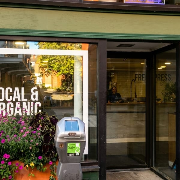 Exterior of a "Local & Organic" shop with a person inside, brick wall, and flower planters.