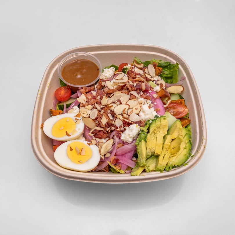 A colorful salad in a brown bowl with sliced avocado, hard-boiled eggs, bacon, almonds, and dressing.
