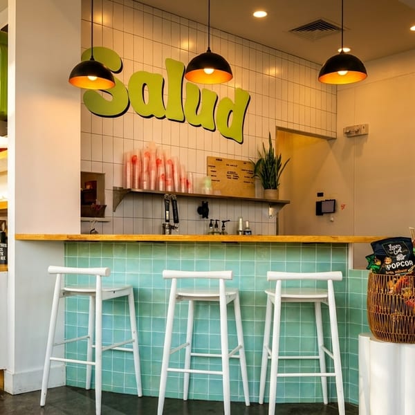 A bright, modern cafe interior with a "Salud" sign, a tiled counter, and white bar stools.