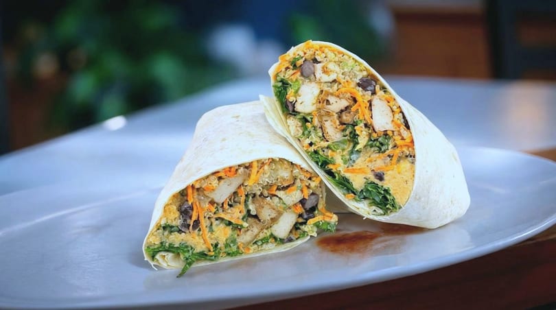 Two halves of a chicken and vegetable wrap, filled with greens, carrots, and beans, on a white plate.