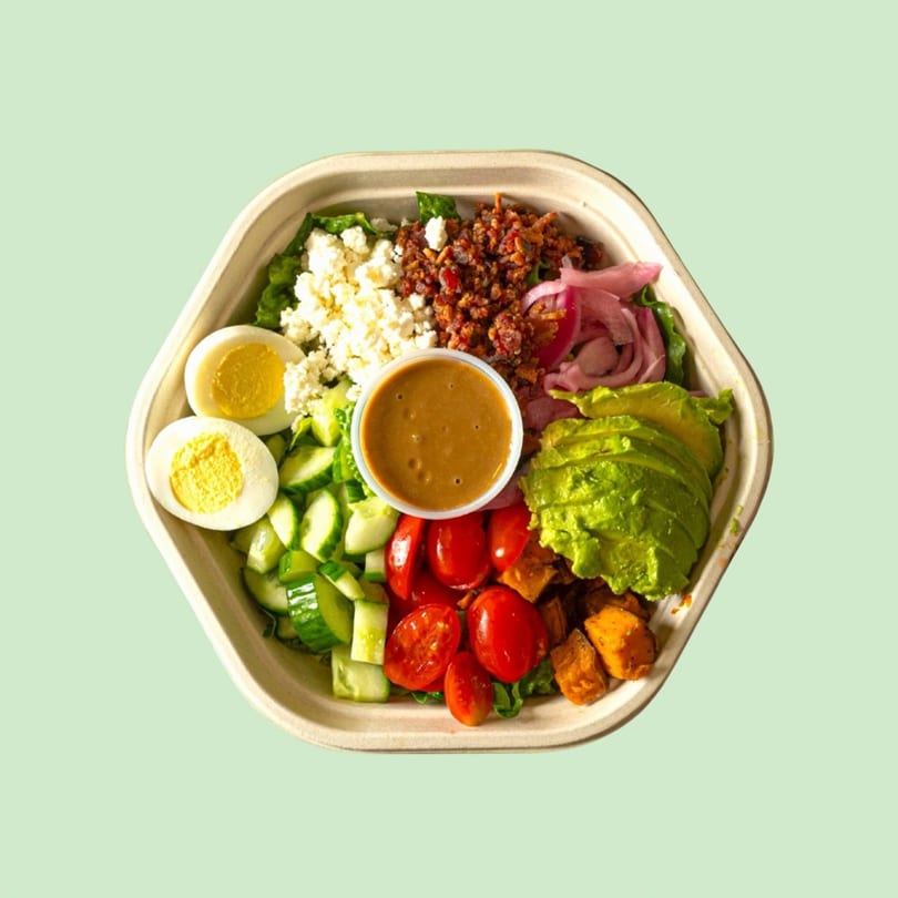 Overhead view of a salad with eggs, bacon, avocado, tomatoes, cucumbers, and dressing in a hexagonal container.