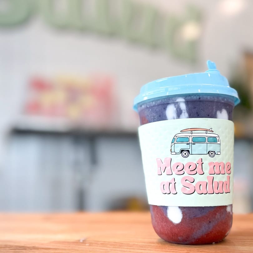 A colorful smoothie with a light blue lid and a "Meet me at Salud" sleeve on a wooden table.